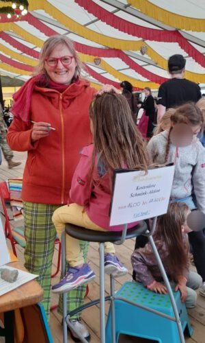 Kinderschminken Hohenfels - deutsch-amerkanisches Freundschaftsfest- Sonja Sonnenschein Frau in rotem Pullover und karierten Hosen kämmt Mädchen mit langen Haaren auf einem hohen Stuhl in einem Festzelt mit rot-gelb-weißen Stoffbahnen an der Decke