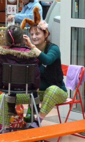 Kinderschminken Ostern - Sonja Sonnenschein Frau mit Hasenohren und karierter Hose schminkt sitzendes Kind mit pinker Haarspange in Innenraum