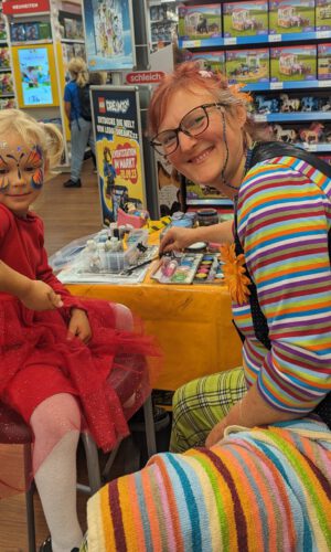 Kind in rotem Kleid sitzt auf Stuhl und lässt sich von Person mit gestreiftem Oberteil und karierten Hosen am Arm bemalen