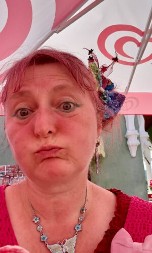 20250813_161150 Frau mit hochgestecktem Haar und buntem Haarschmuck trägt eine Halskette mit Schmetterlingsanhänger und sitzt unter einem Sonnenschirm mit roten Spiralmustern