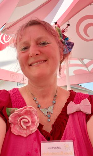 Frau mit rosa Kleid und rotem Oberteil trägt eine Halskette mit Schmetterlingsanhänger und eine große Rosenbrosche am Kleid unter einem Sonnenschirm mit Herzmotiven