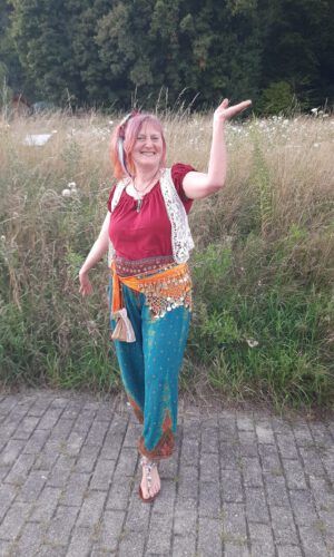 Person mit buntem, folkloristischem Kleidungsstil steht barfuß auf gepflastertem Weg vor hohem Gras und hebt eine Hand nach oben