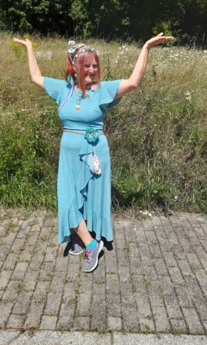 Person in blauem Kleid mit Blumenmuster und Sportschuhen steht auf gepflastertem Weg vor hohem Gras und hebt beide Arme seitlich nach oben