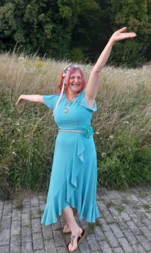 Frau in blauem Kleid mit ausgestreckten Armen steht auf gepflastertem Weg vor hohem Gras und Bäumen