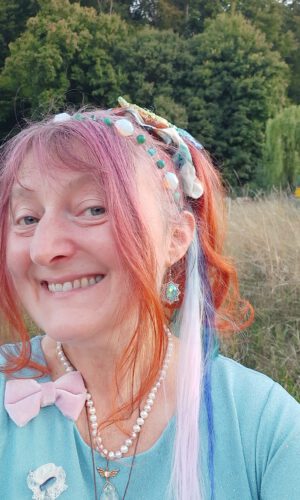 Person mit langem, teils geflochtenem Haar in verschiedenen Farben, trägt eine Perlenkette, einen rosa Schleifenbrosche und bunte Ohrringe vor einer Wiese und Bäumen