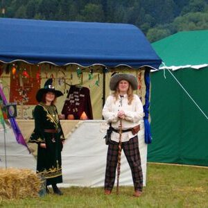 Zwei Personen in historischer Kleidung stehen vor einem Marktstand mit blauer Plane und daneben einem grünen Zelt auf einer Wiese
