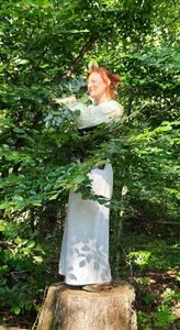 Frau in weißem Kleid steht auf einem Baumstumpf umgeben von dichtem Laubwerk