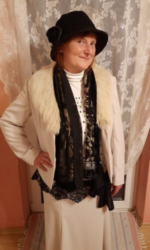 Person steht vor einem weißen Spitzenvorhang trägt schwarzen Hut weißen Mantel mit Fellkragen schwarzen Schal und weißen Rock