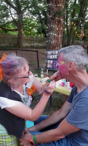 Facepainting medbo Frau mit lila Haaren und orangefarbener Blume im Haar malt einem sitzenden Mann mit grauen Haaren im Freien das Gesicht, im Hintergrund Bäume und eine Holzzaun.