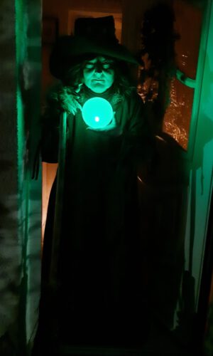 Person mit breitkrempigem Hut und Umhang hält leuchtende Kugel in dunklem Raum