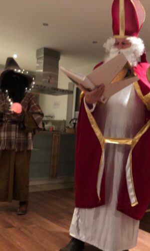 Zwei Personen in einem Raum, eine als Nikolaus mit Bischofsmütze, rotem Umhang und Bischofsstab, die andere in dunkler Verkleidung mit Kapuze und kariertem Umhang