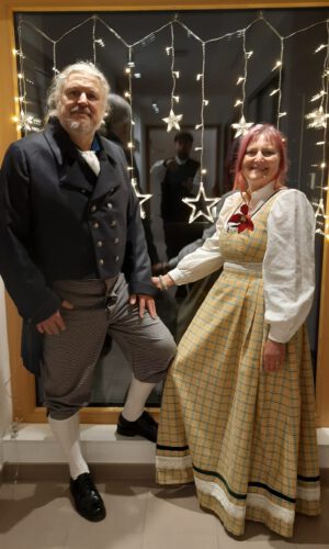 Zwei Personen in historischer Kleidung vor einem Fenster mit Sternenlichterkette, eine Person trägt Kniebundhose und Gehrock, die andere ein langes Kleid mit Puffärmeln