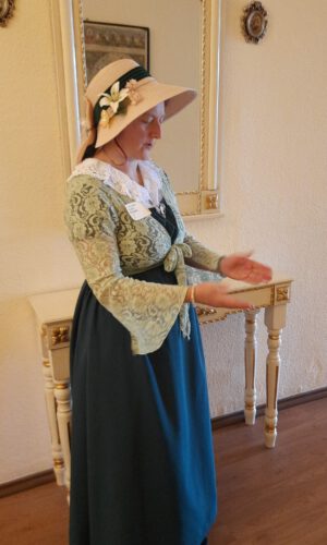 Frau in historischer Tracht mit Hut steht seitlich vor einem Spiegel und einem kleinen Tisch mit verzierten Beinen