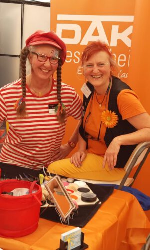 Schminkstand_Kinderschminken Zwei Personen sitzen an einem orange gedeckten Tisch mit verschiedenen Utensilien vor orangefarbenen Wänden mit dem Schriftzug 'DAK Gesundheit'