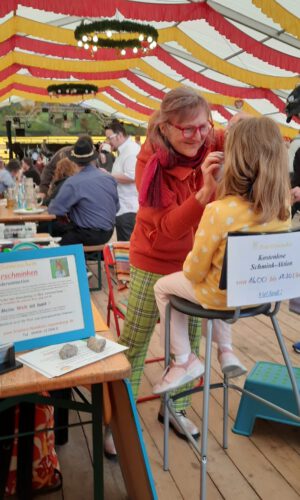 Sonja Sonnenschein Kinderschminken Hohenfels Zwei Frauen in einem Festzelt mit rot-gelben Stoffbahnen an der Decke, eine Frau trägt ein rotes Oberteil und karierte Hose und schminkt die andere Frau, die auf einem hohen Stuhl sitzt.