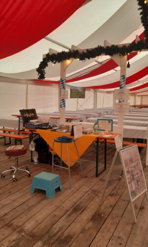 Volksfest Beratzhausen Kinderschminken Kinderschmink-Stand auf einem Festival: Unter einem Zelt stehen Tische und Bänke, ein Schild mit der Aufschrift ‚Kinderschminken‘ ist sichtbar.