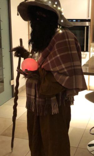 Person mit langem Bart und spitzem Hut, der mit Lichterkette geschmückt ist, hält einen leuchtenden Ball und einen gedrehten Stab