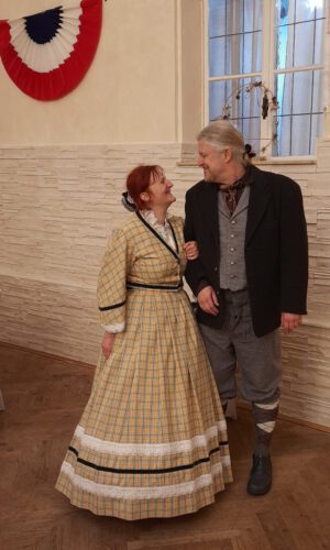 Zwei Personen in einem Raum, eine als Nikolaus mit Bischofsmütze, rotem Umhang und Bischofsstab, die andere in dunkler Verkleidung mit Kapuze und kariertem Umhang