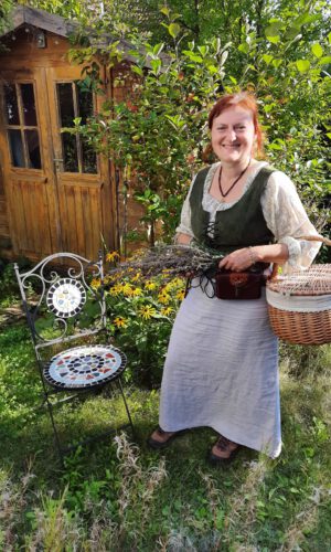 Frau in traditioneller Kleidung mit Korb und Bündel Kräuter steht neben einem Stuhl mit Mosaikmuster vor einem Holzhaus im Garten