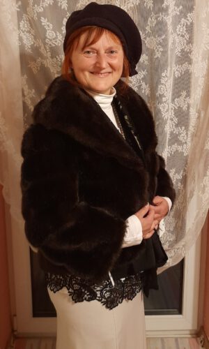 Person mit schwarzer Baskenmütze, schwarzem Pelzmantel und weißem Rollkragenpullover vor weißer Gardine