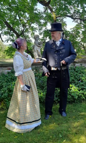 Ein Mann in dunklem Anzug mit Zylinder und Gehstock steht neben einer Frau in traditionellem kariertem Kleid mit weißer Bluse und Handschuhen vor einer Steinmauer und einer Statue im Garten