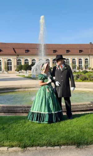 Ein Paar in historischer Kleidung steht vor einem runden Springbrunnen mit Fontäne auf einer Rasenfläche vor einem langgestreckten Gebäude mit vielen Fenstern