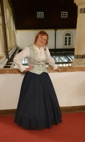 Frau in historischer Tracht mit weißer Bluse, gemusterter Weste und langem dunklem Rock lehnt an Brüstung in einem Innenhof mit Säulen und Fenstern